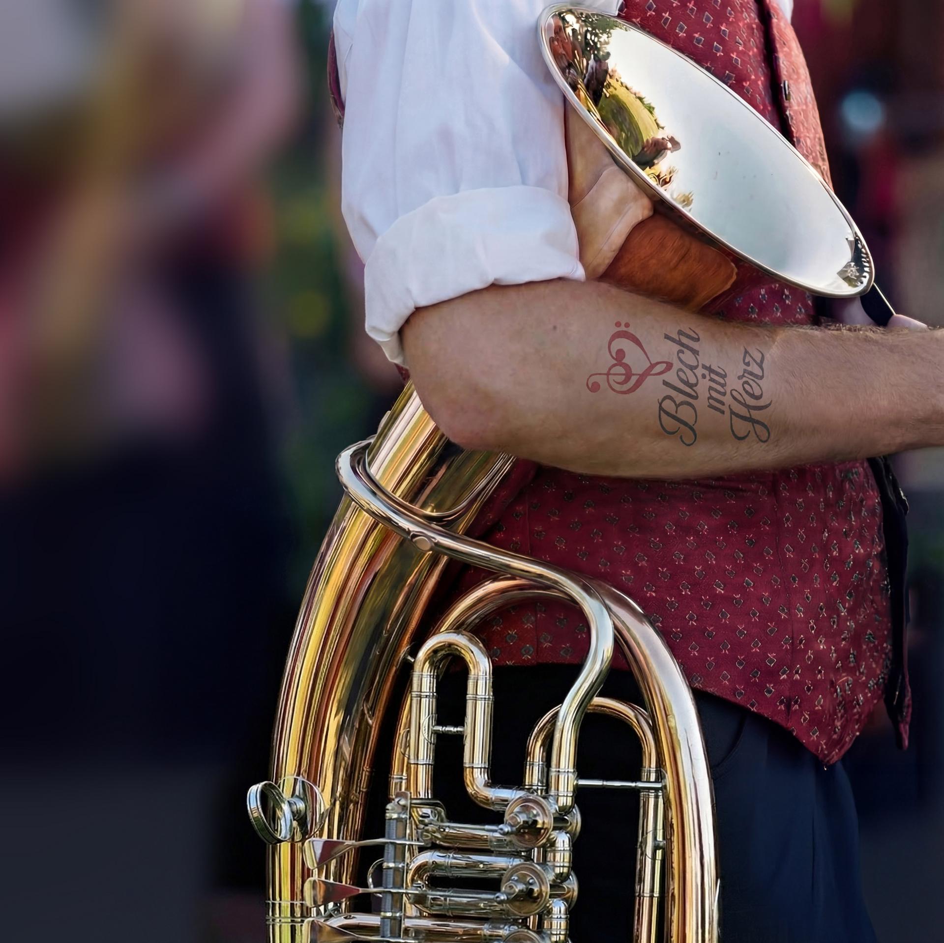 Plakat mit Nahaufnahme einer Person mit Blechblasinstrument und Tatoo auf dem Unterarm mit dem Text „Blech mit Herz“