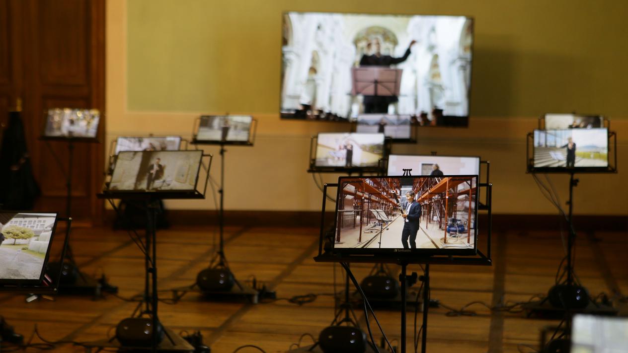 Medieninstallation in einem Ausstellungsraum. An 22 Notenpulte sind Monitore angebracht, die Videos von einzelnen Musiker:innen zeigen.