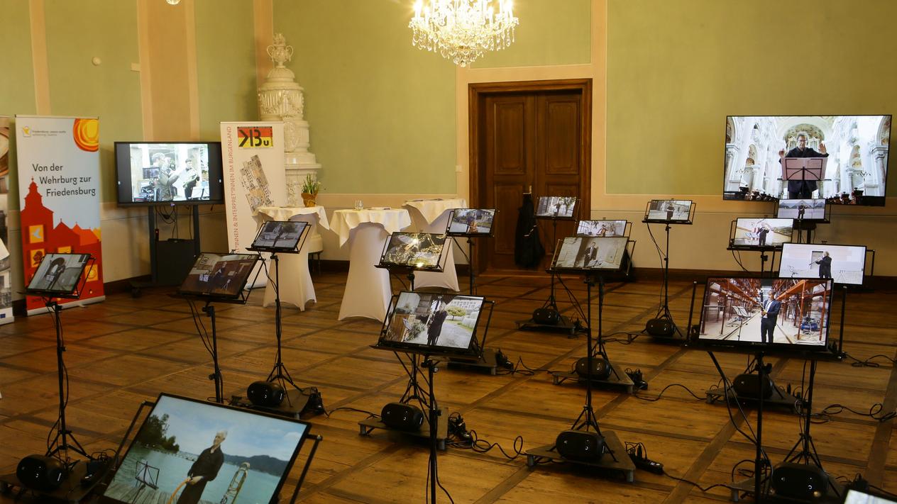Medieninstallation in einem Ausstellungsraum. An 22 Notenpulte sind Monitore angebracht, die Videos von einzelnen Musiker:innen zeigen.