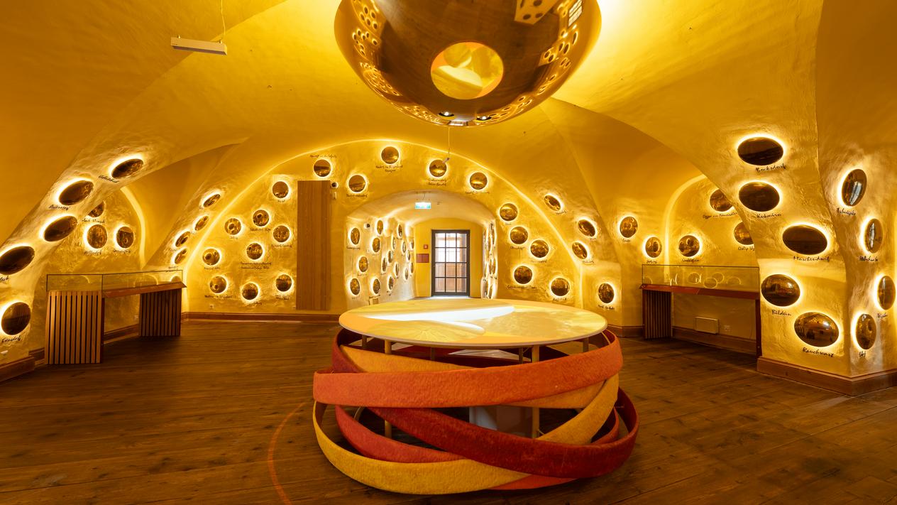 Ein Ausstellungsraum mit einem runden Tisch und beleuchteten Scheiben an den Wänden, die verschiedene Ortschaften symbolisieren